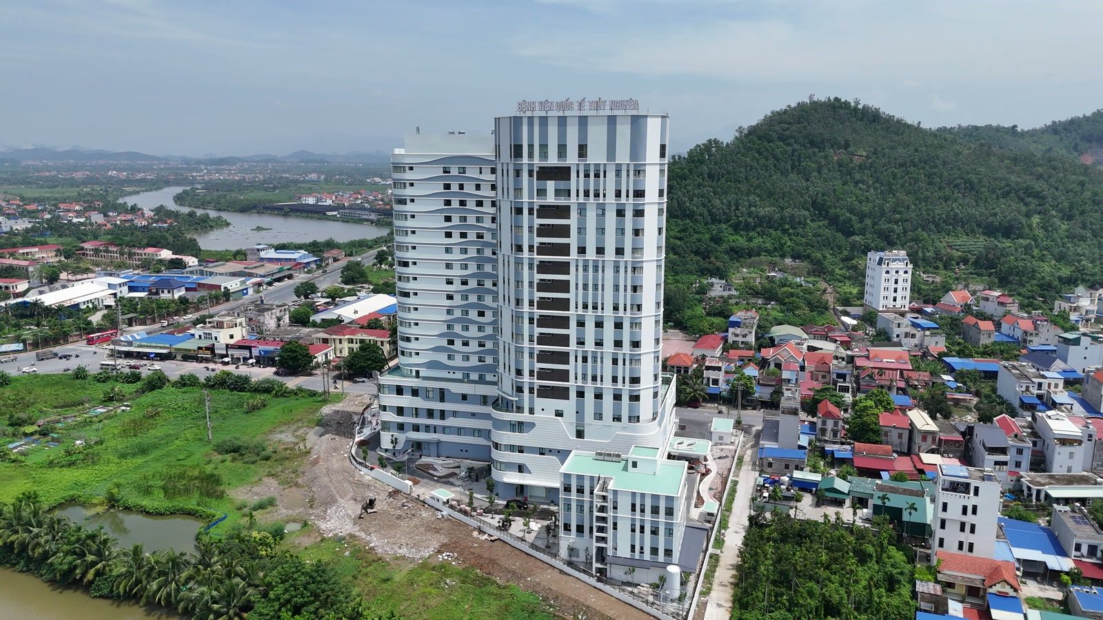 Toà Bệnh viện Đa khoa Quốc tế Thủy Nguyên 19 tầng đứng một mình một khối giữa khu dân cư mái đỏ, phía sau là núi, nhìn từ flycam