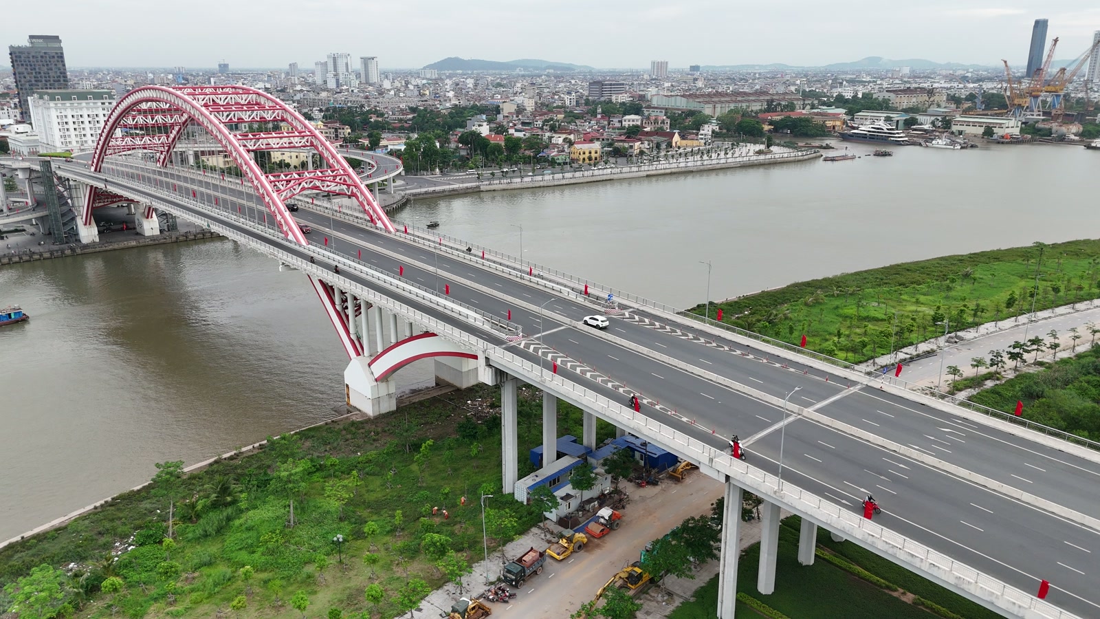 Cầu Hoàng Văn Thụ vòm thép đỏ bắc qua sông Cấm sang Bắc Sông Cấm, phần đường dẫn nhiều làn chạy xuống bãi cỏ xanh ở bờ bắc, phía xa bên kia sông là nội đô Hải Phòng