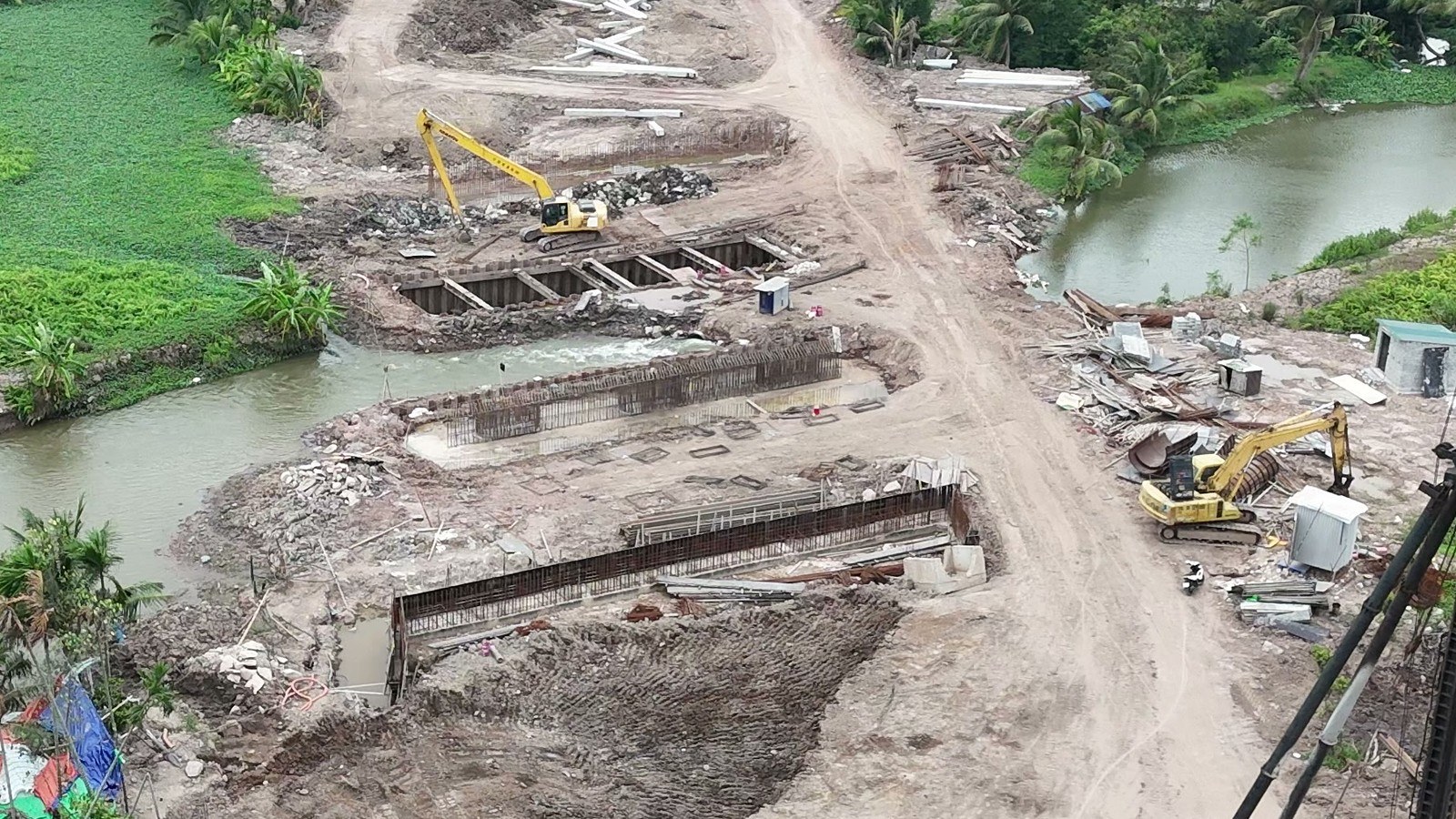 Hố móng cầu qua kênh trên đường 2,4km tháng 9/2026, trong hố bên này bờ kênh đã dựng những khung cốt thép dài xếp thẳng hàng, hố phía xa vẫn còn quây cừ thép, máy xúc đứng bên cạnh