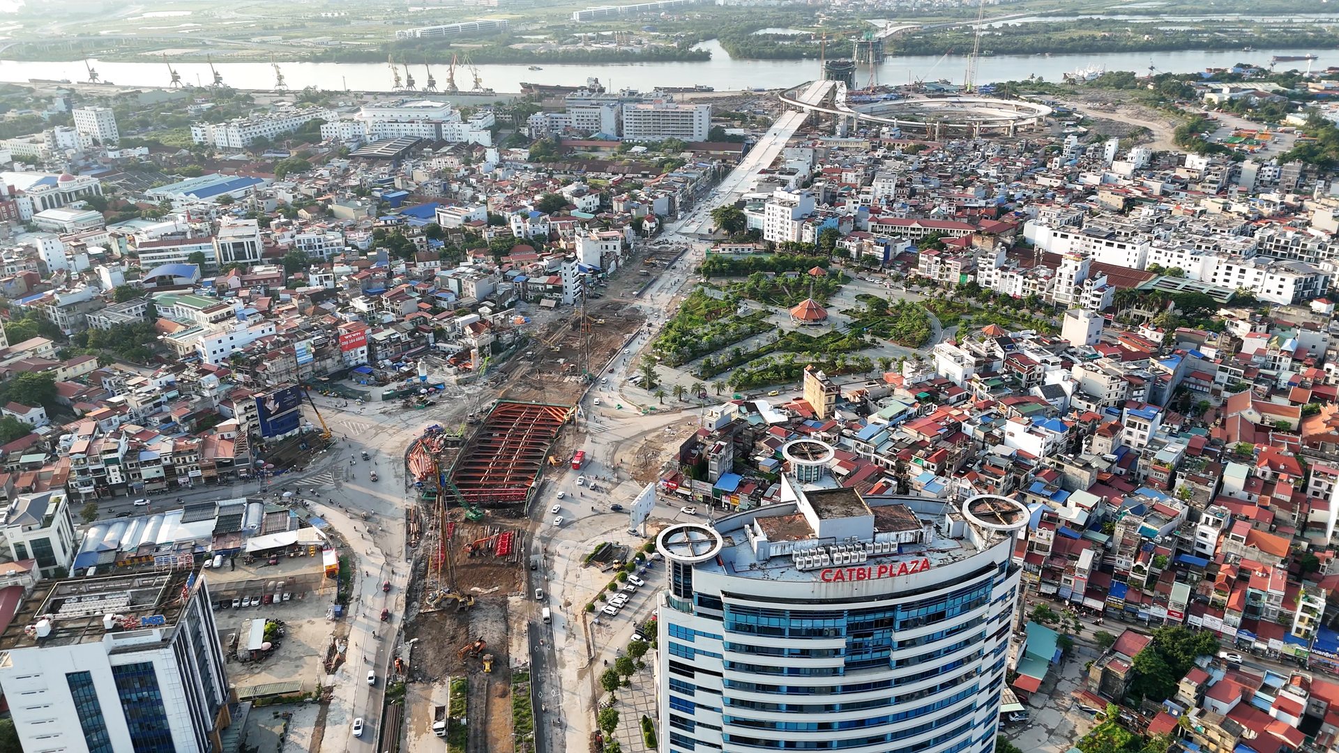 Công trường hầm chui Ngã 6 Lạc Viên với khung thép đỏ chống miệng hố, phía trước là toà Cát Bi Plaza, phía xa là sông Cấm