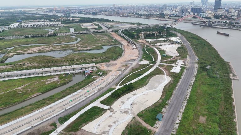 Dải công viên ven sông Cấm đang làm dở chạy dọc bờ sông, bên trái là con kênh phía trong, bên phải là đường ven sông, phía xa là cầu vòm đỏ Hoàng Văn Thụ và nội đô Hải Phòng