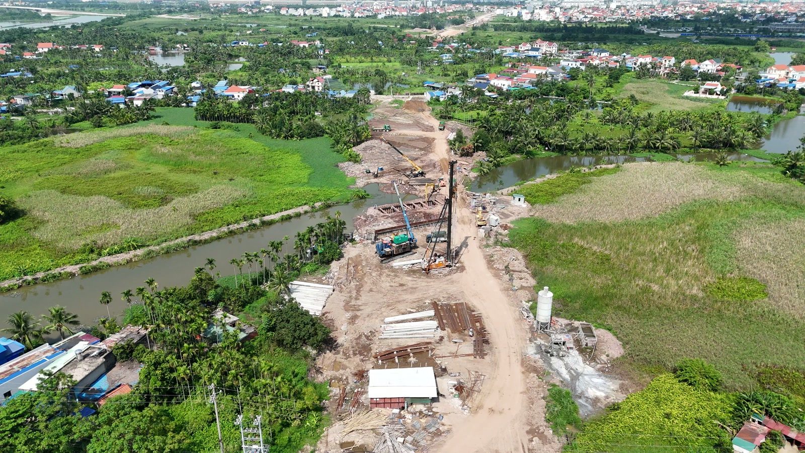 Công trường cầu qua kênh trên tuyến đường 2,4km, máy đóng cọc dựng cao bên mép nước, vật liệu tập kết dọc đường