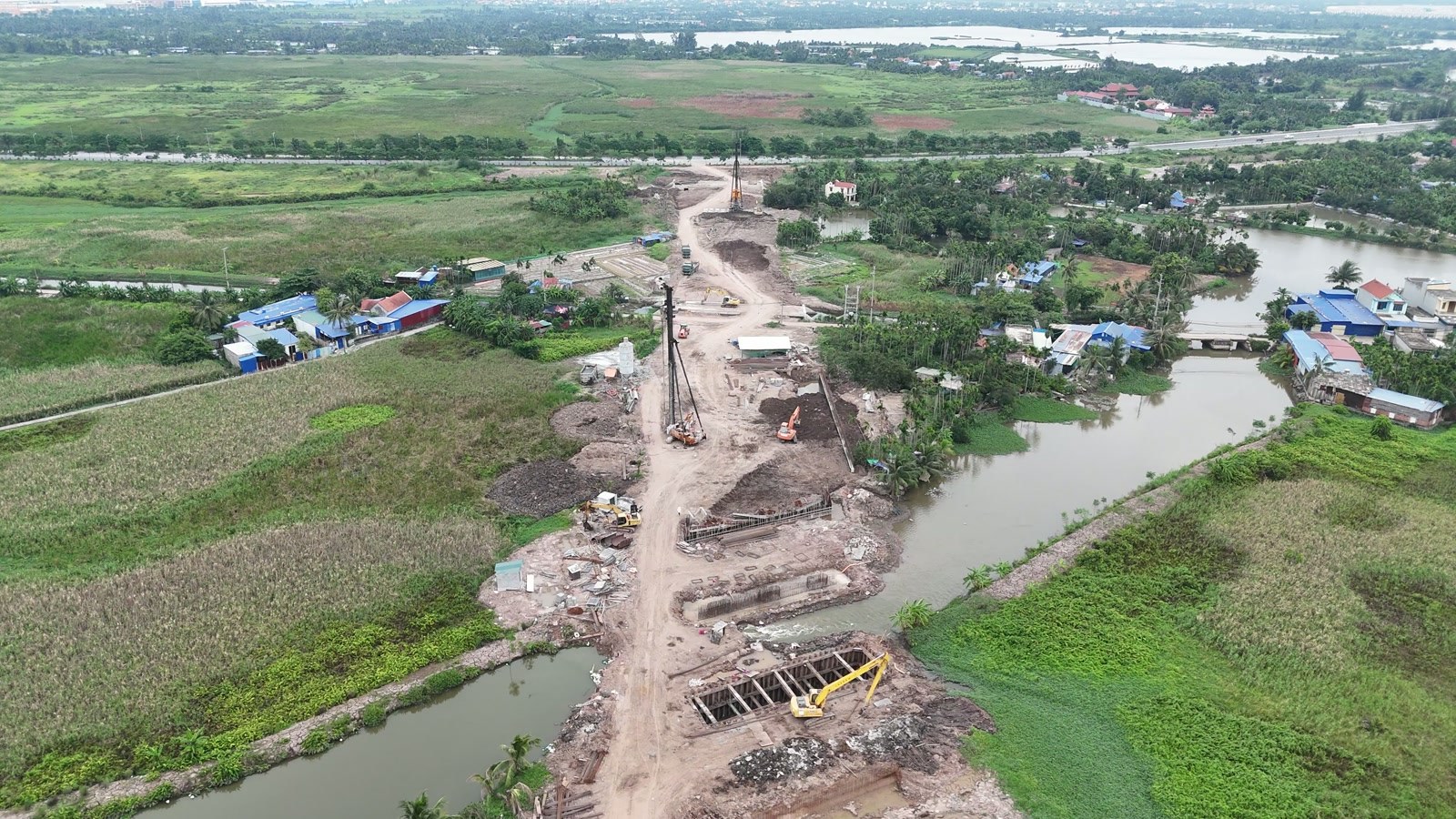 Chỗ đường 2,4km cắt ngang con kênh: máy khoan cọc xi măng đất, hố móng cầu quây cừ thép hai bên bờ, xung quanh là ruộng và làng xóm