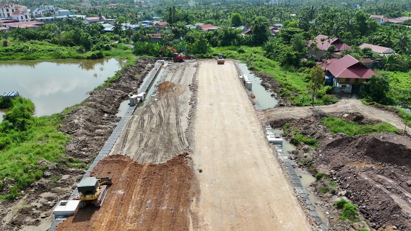 Nền đường 2,4km đắp cao chạy giữa hai hồ nước, hai bên mép đã kè bằng tấm bê tông đúc sẵn, máy ủi đang san mặt nền