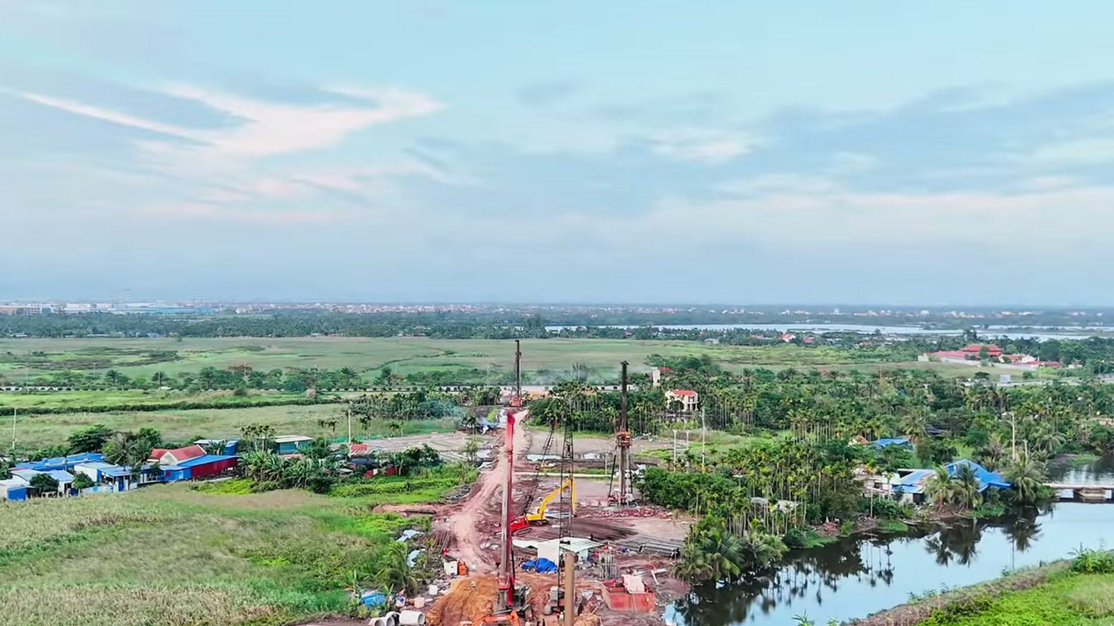 Công trường bên mép kênh với máy khoan cọc dựng cao, nền đất đỏ và vật liệu, bên cạnh là ruộng và nhà dân