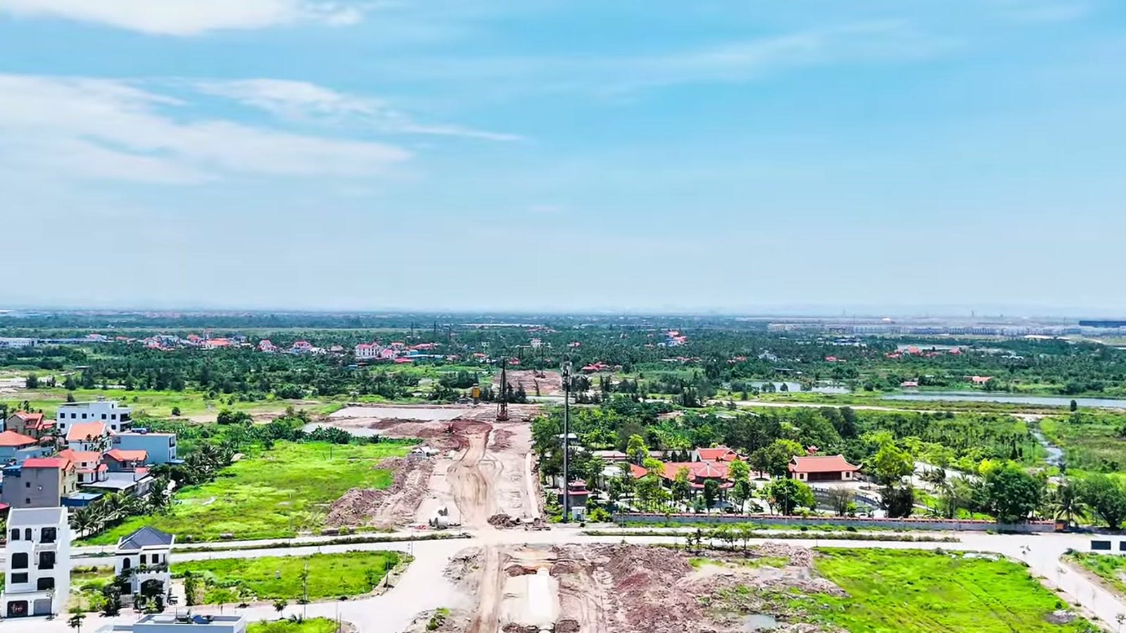 Khoảng đất đang san nền nằm giữa các dãy nhà dân và bãi cỏ, mặt đất còn vết máy, phía xa là ruộng và hàng cây
