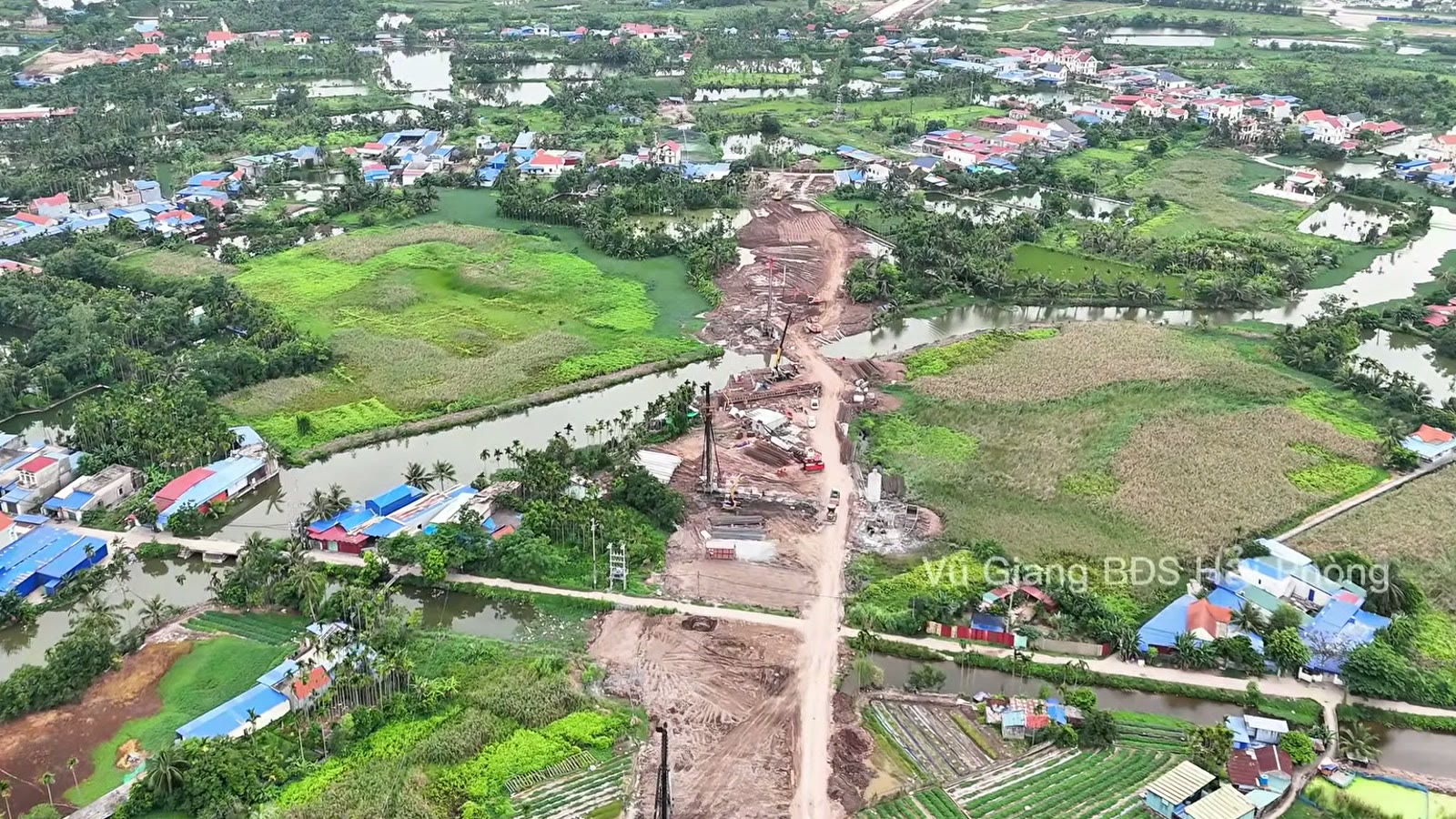 Dải nền đường đất đỏ chạy qua vùng trũng có nhiều ao và mương, hai bên là cây xanh và mái nhà dân