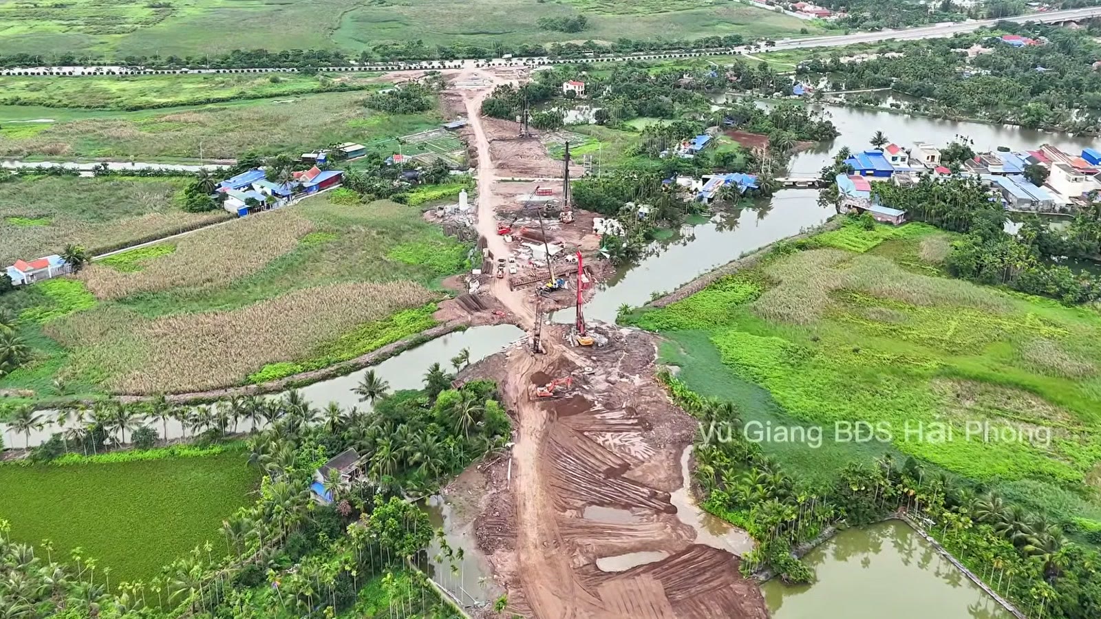 Nền đường đất đỏ đi qua một con kênh giữa ruộng, có máy khoan cọc đứng trên nền, hai bên là ao và nhà dân