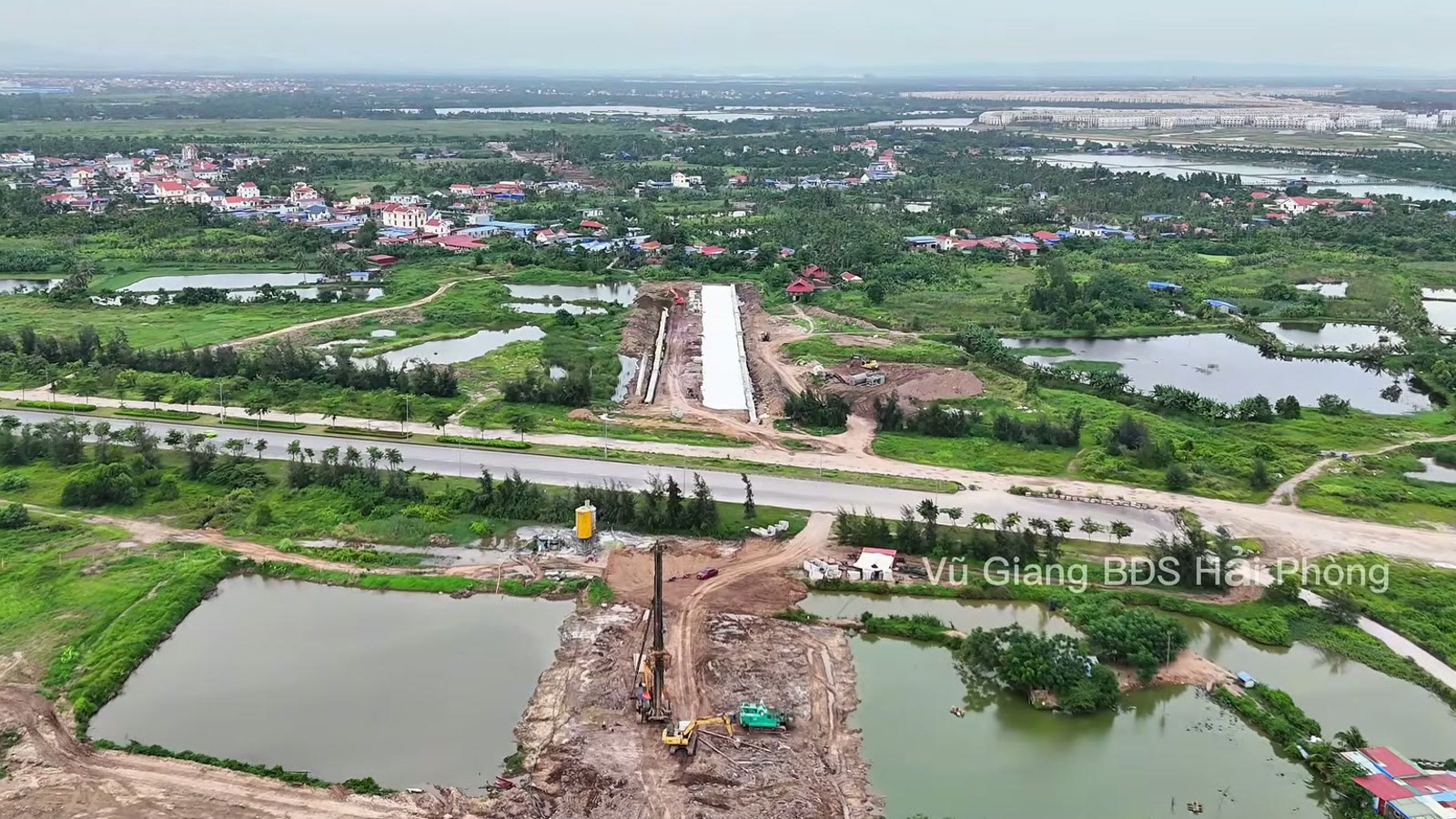 Máy khoan cọc làm giữa khu ao sát đại lộ Bắc Nam VSIP đã trải nhựa, bên kia đại lộ là nền đường có hàng cấu kiện bê tông trắng, xa hơn là ruộng và làng