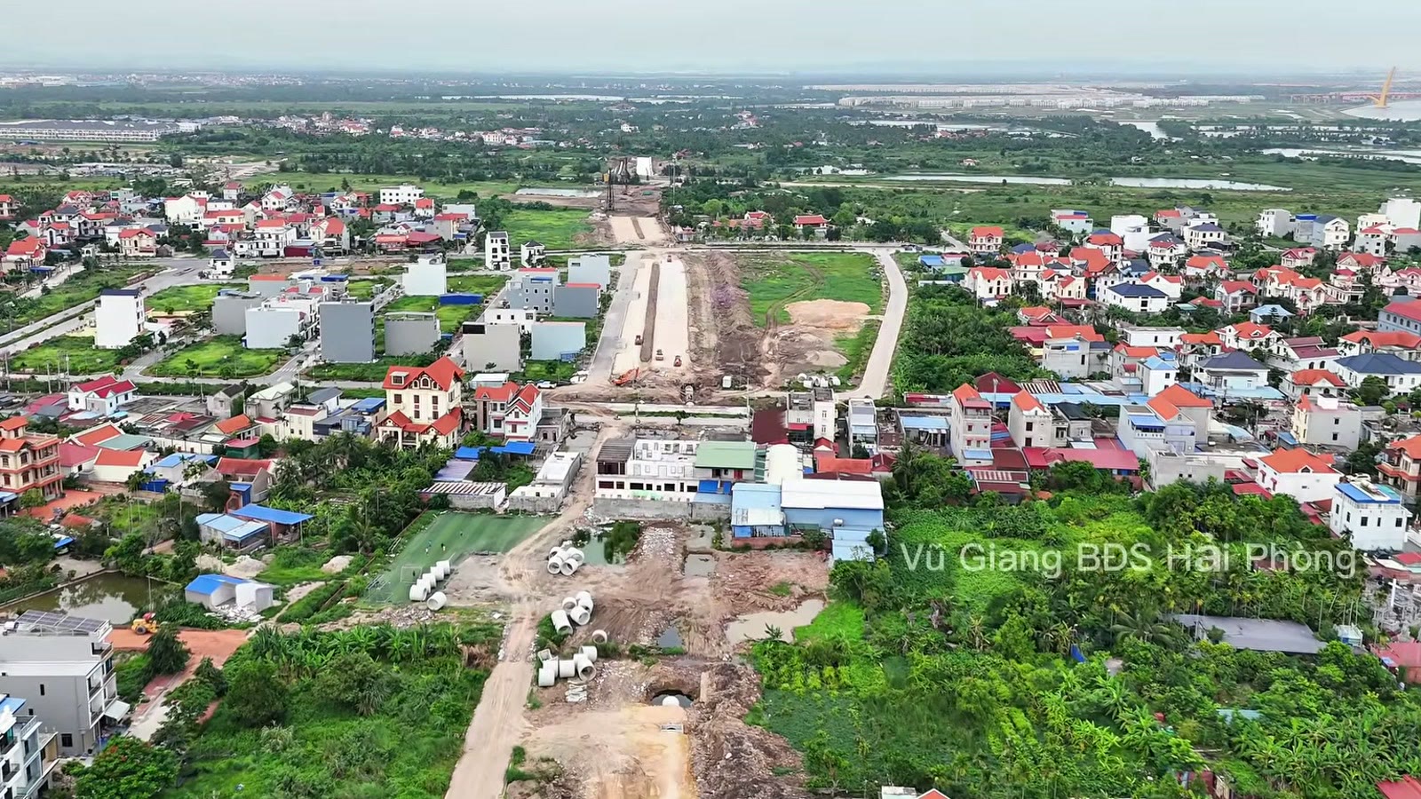 Nền đường đất chạy qua khu dân cư đông nhà, hai bên là nhà ở nhiều tầng và vườn cây, trên nền còn vật liệu và cống bê tông
