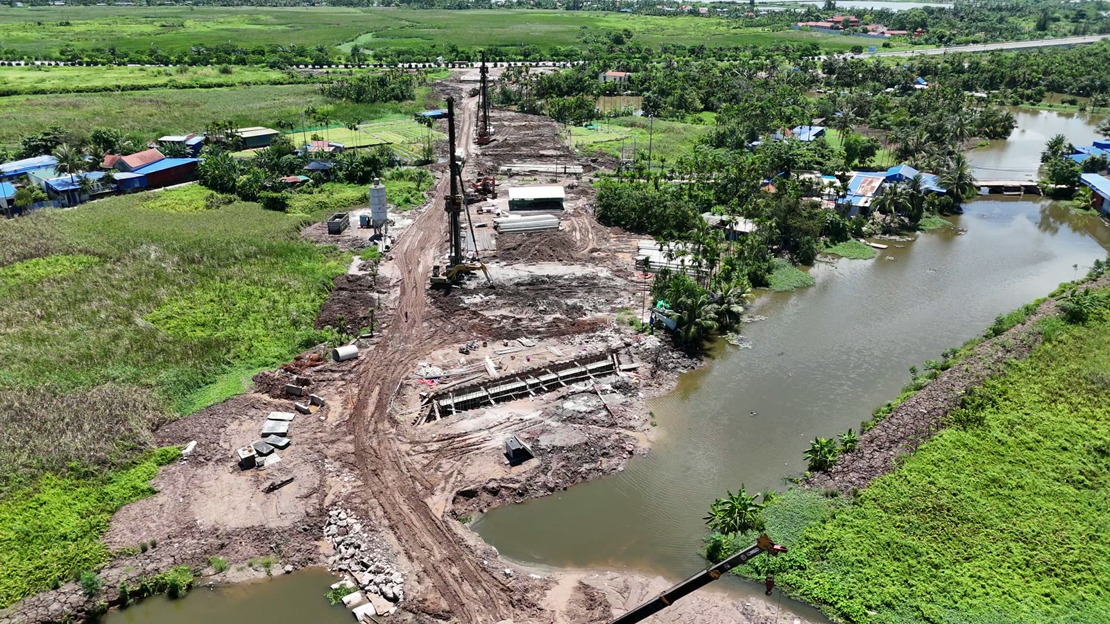 Máy khoan cọc dựng cao bên mép một con kênh, trên nền đất có cấu kiện bê tông và vật liệu, hai bên là ruộng và cây xanh