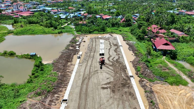 Nền đường đất rộng với hai hàng cấu kiện bê tông thoát nước dọc hai bên và máy xúc đang làm, đường 2,4km Đỗ Mười ra đảo Vũ Yên ngày 12/7/2026