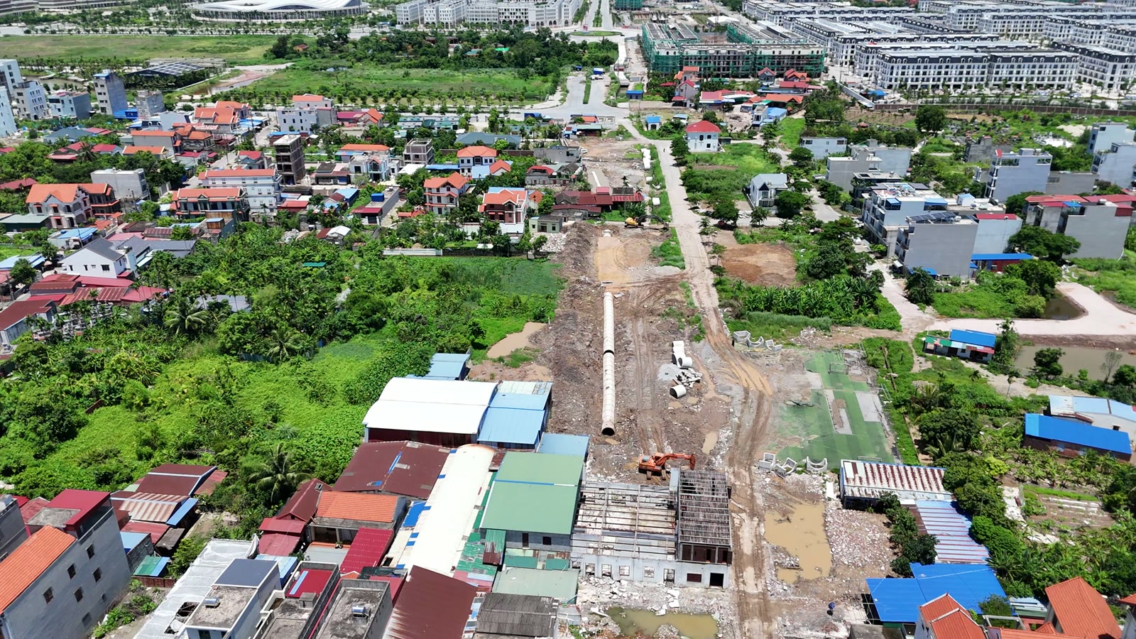 Nền đường đất chạy xuyên qua khu dân cư đông nhà, trên nền có ống cống bê tông nằm dọc, phía xa là các dãy nhà phố mới xây