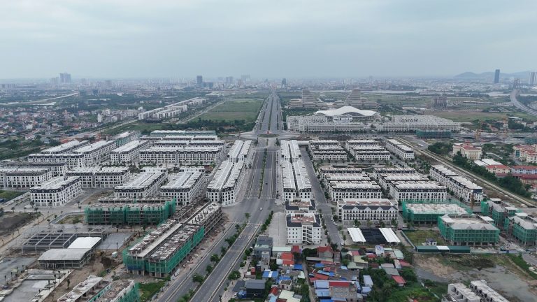 Toàn cảnh Hoàng Huy New City tháng 9/2026 nhìn dọc đường Đỗ Mười ở Bắc Sông Cấm, các dãy nhà thấp tầng mái đen trắng xếp thành ô hai bên trục đường, cuối trục là khu trung tâm hành chính mới và nội đô Hải Phòng bên kia sông