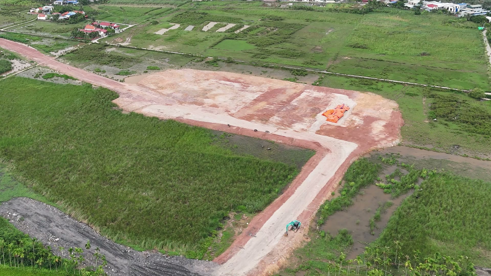 Mảng đất đã san nền màu nâu đỏ nối với con đường đất mới đắp, trên đường có một chiếc máy xúc, xung quanh là ruộng xanh