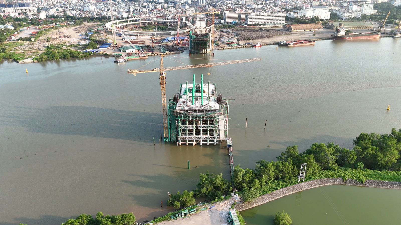 Trụ tháp cầu Nguyễn Trãi đang xây nhô lên giữa lòng sông Cấm, có cần cẩu tháp màu vàng bên cạnh, hai bên là bờ cây xanh và công trường ở bờ nội đô