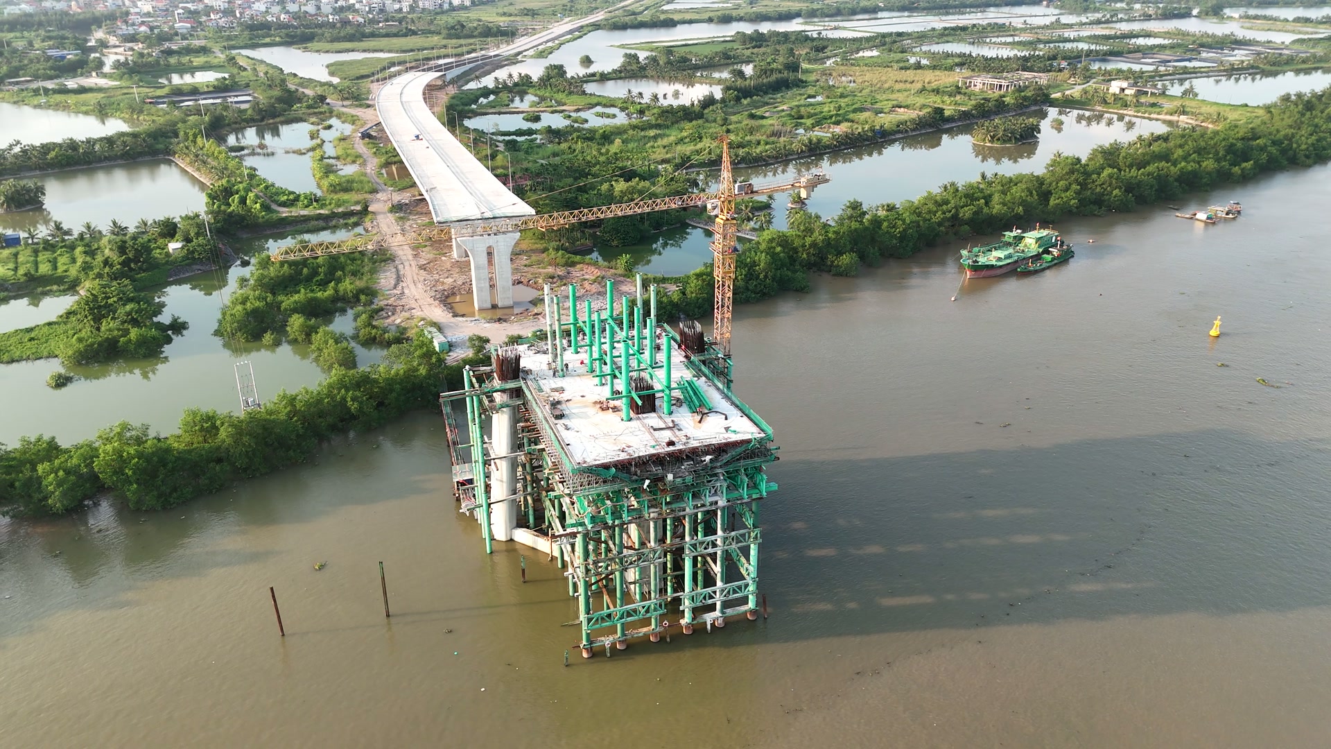 Trụ tháp cầu Nguyễn Trãi phía Thủy Nguyên với mặt bệ đã đổ bê tông xong, trên đó dựng thêm một tầng giàn giáo xanh và các bó cốt thép chờ, cầu dẫn màu trắng chạy tới mép sông