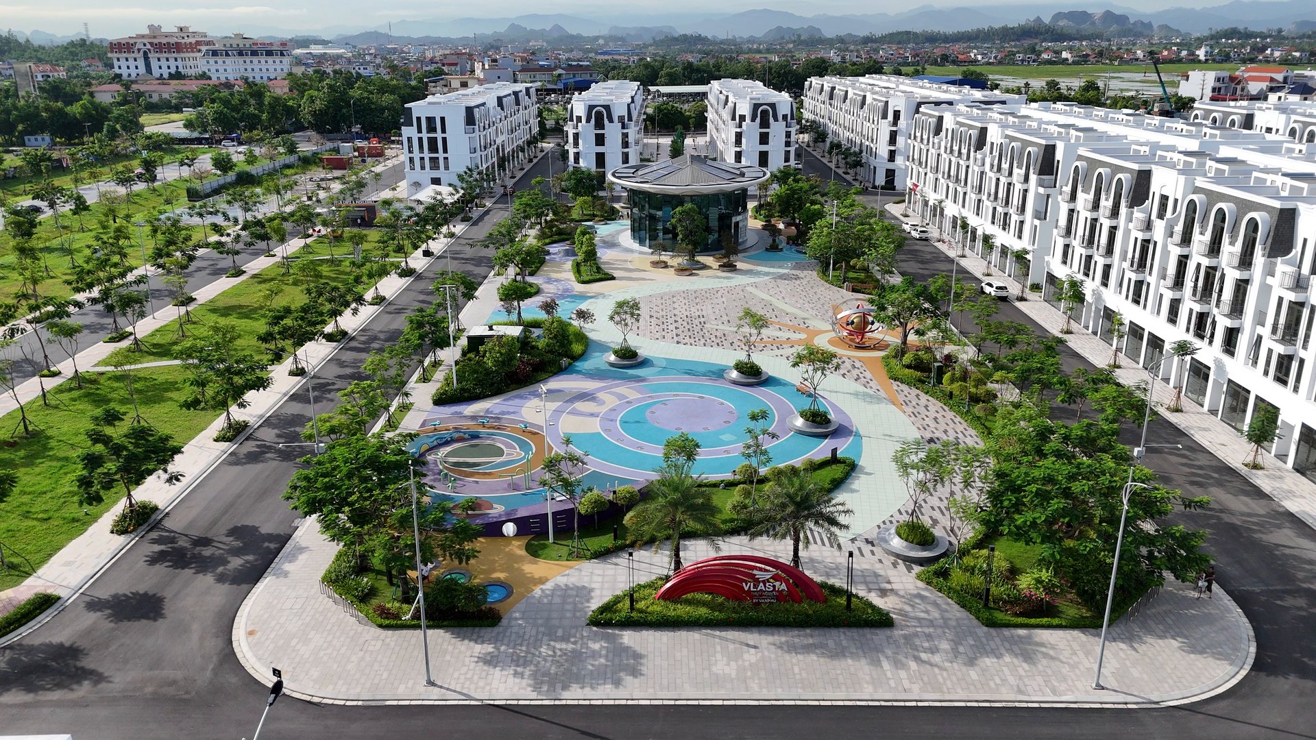 Quảng trường sân chơi trẻ em lát gạch màu trong khu đô thị Vlasta Hải Phòng, xung quanh là cây xanh và những dãy nhà thấp tầng đã xây xong