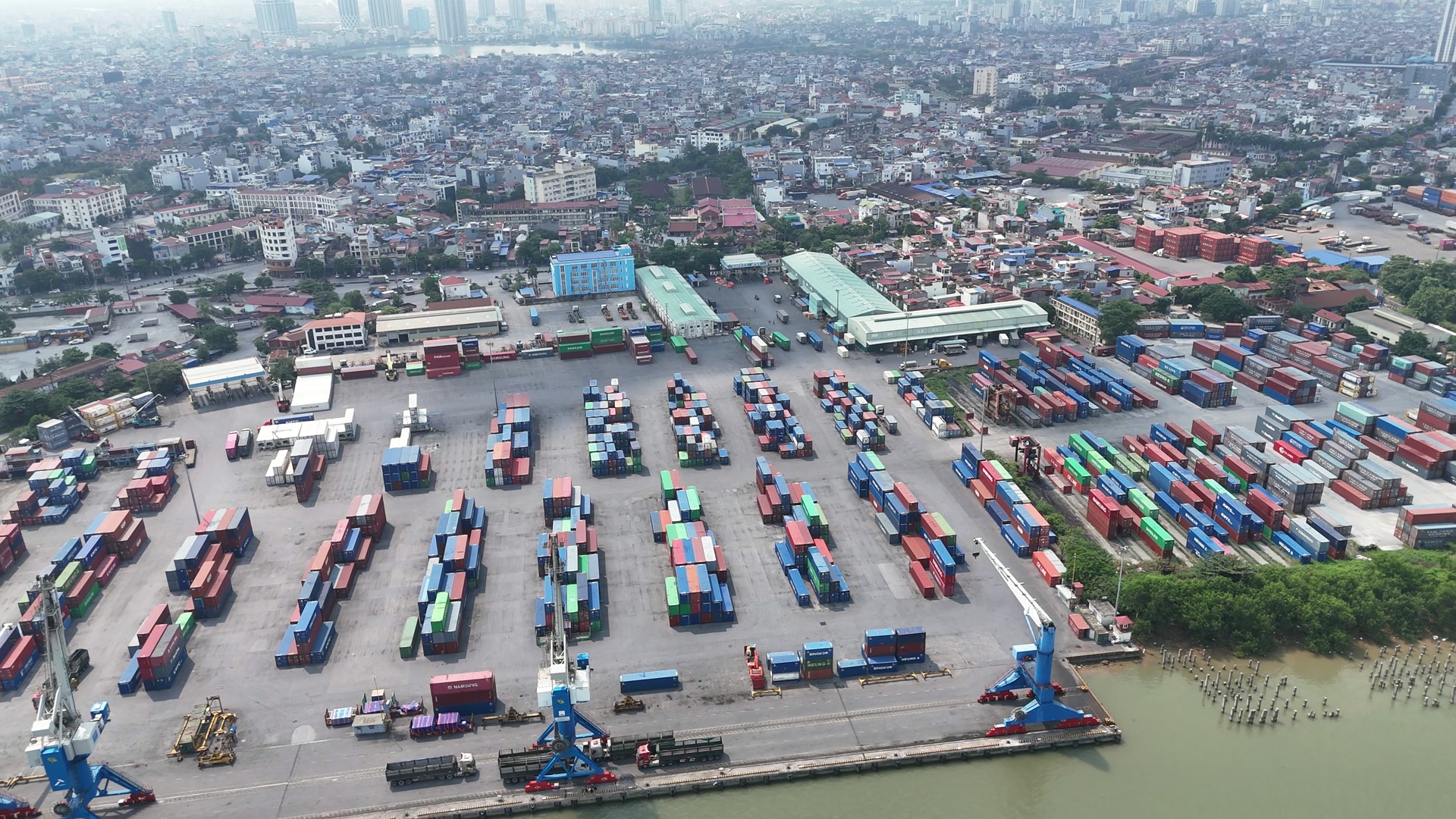 Bãi container nhiều màu xếp thành từng lô trên mặt bến bê tông, hai cần cẩu chân đế màu xanh và dãy xe đầu kéo ở mép bến sát sông, phía sau là nhà xưởng mái xanh và khu dân cư nhà ống san sát