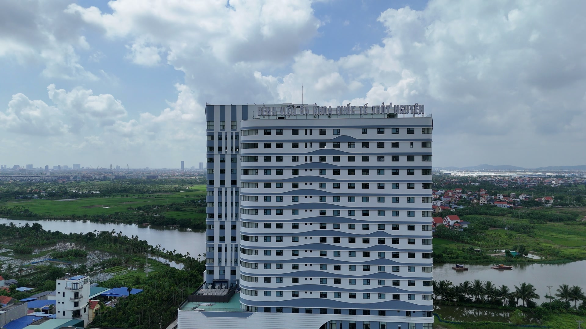 Toà nhà 19 tầng của Bệnh viện Đa khoa Quốc tế Thủy Nguyên với dòng chữ tên bệnh viện gắn trên mái, phía sau là sông và đồng ruộng