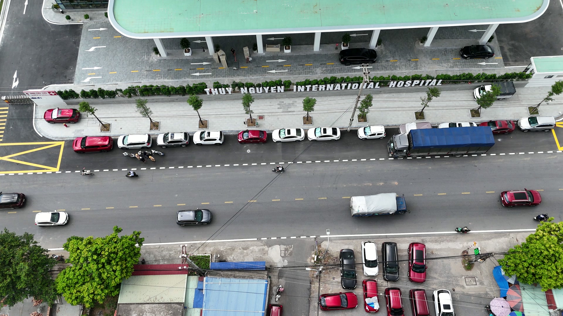 Hàng ô tô đỗ dọc mép đường trước Bệnh viện Đa khoa Quốc tế Thủy Nguyên, xe tải và xe máy qua lại trên tuyến đường chạy trước bệnh viện