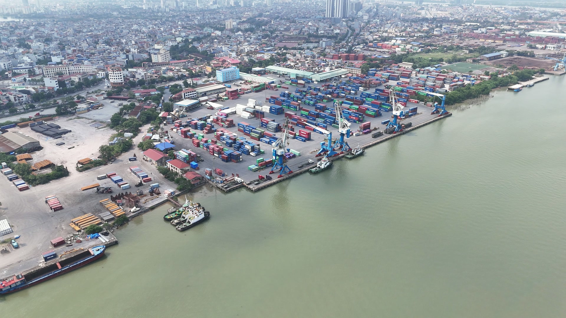Góc một dải bến trên sông Cấm, bãi container xếp từng lô, mấy cần cẩu chân đế màu xanh đứng dọc mép bến, mấy chiếc tàu kéo đậu sát bờ, góc dưới là một sà lan, phía sau là khu dân cư dày đặc