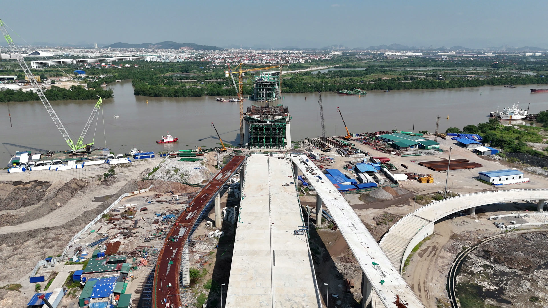 Trụ tháp cầu Nguyễn Trãi phía nội đô, khối bê tông quây giàn giáo xanh sát bờ sông Cấm, trên bờ là bãi thép và ống thép xếp thành đống, cần cẩu đứng cạnh