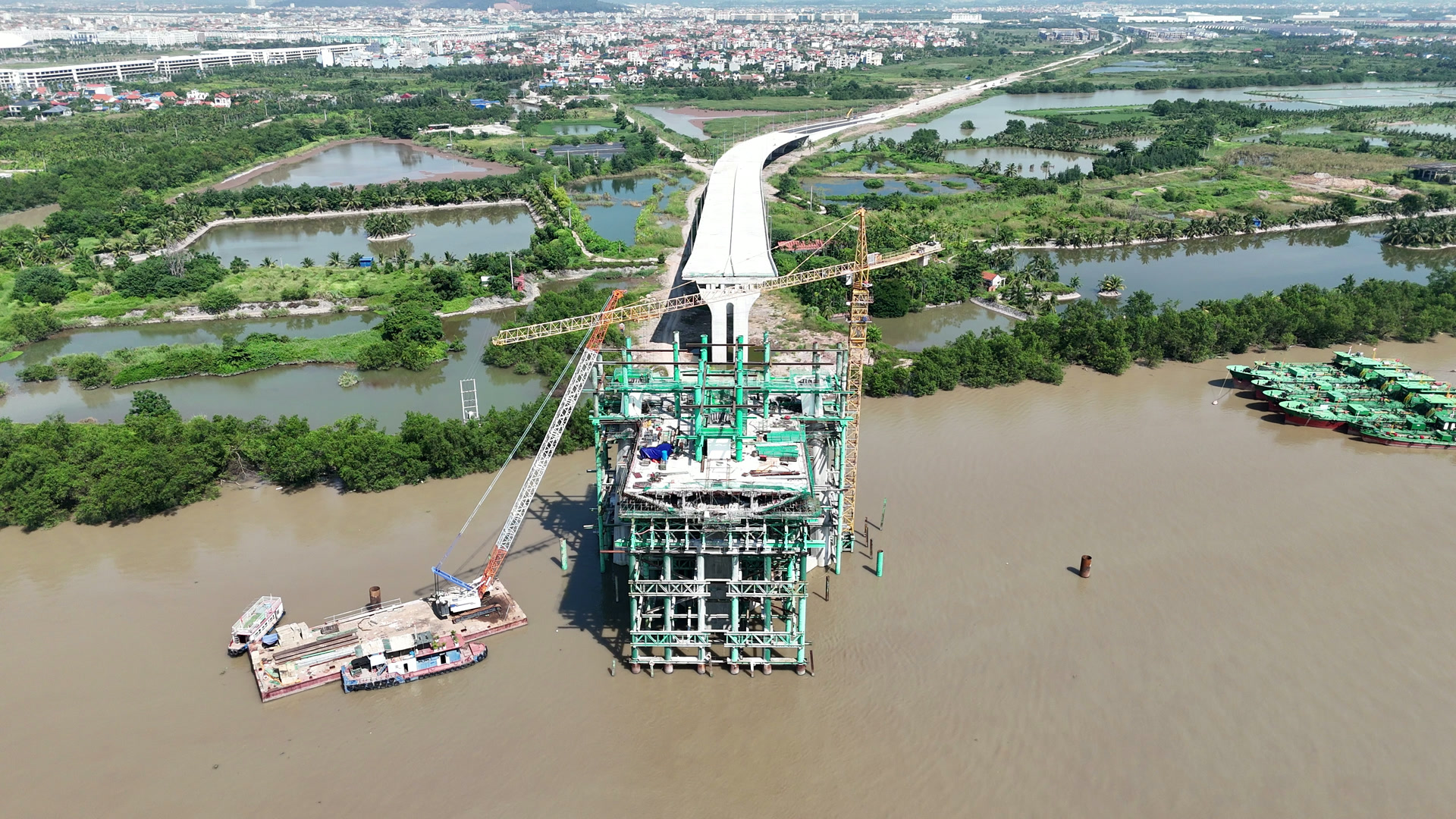 Trụ tháp cầu Nguyễn Trãi phía Thủy Nguyên giữa sông Cấm, khung giàn giáo xanh nhiều tầng, cần cẩu tháp màu vàng dựng trên trụ, cầu dẫn bê tông trắng phía sau chạy vào bờ