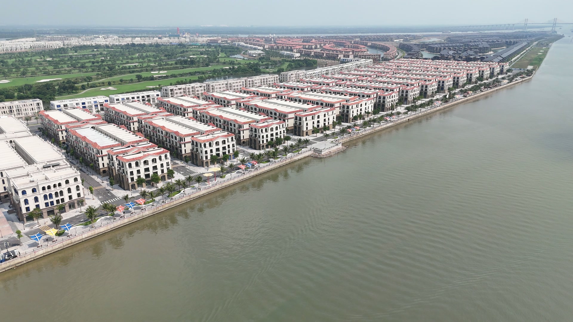 Dãy nhà phố viền ngói đỏ quanh mép mái chạy dài dọc bờ sông Cấm trên đảo Vũ Yên, sát mép nước là phố đi bộ lát đá có hàng dừa và các ô dù màu, phía sau là sân golf