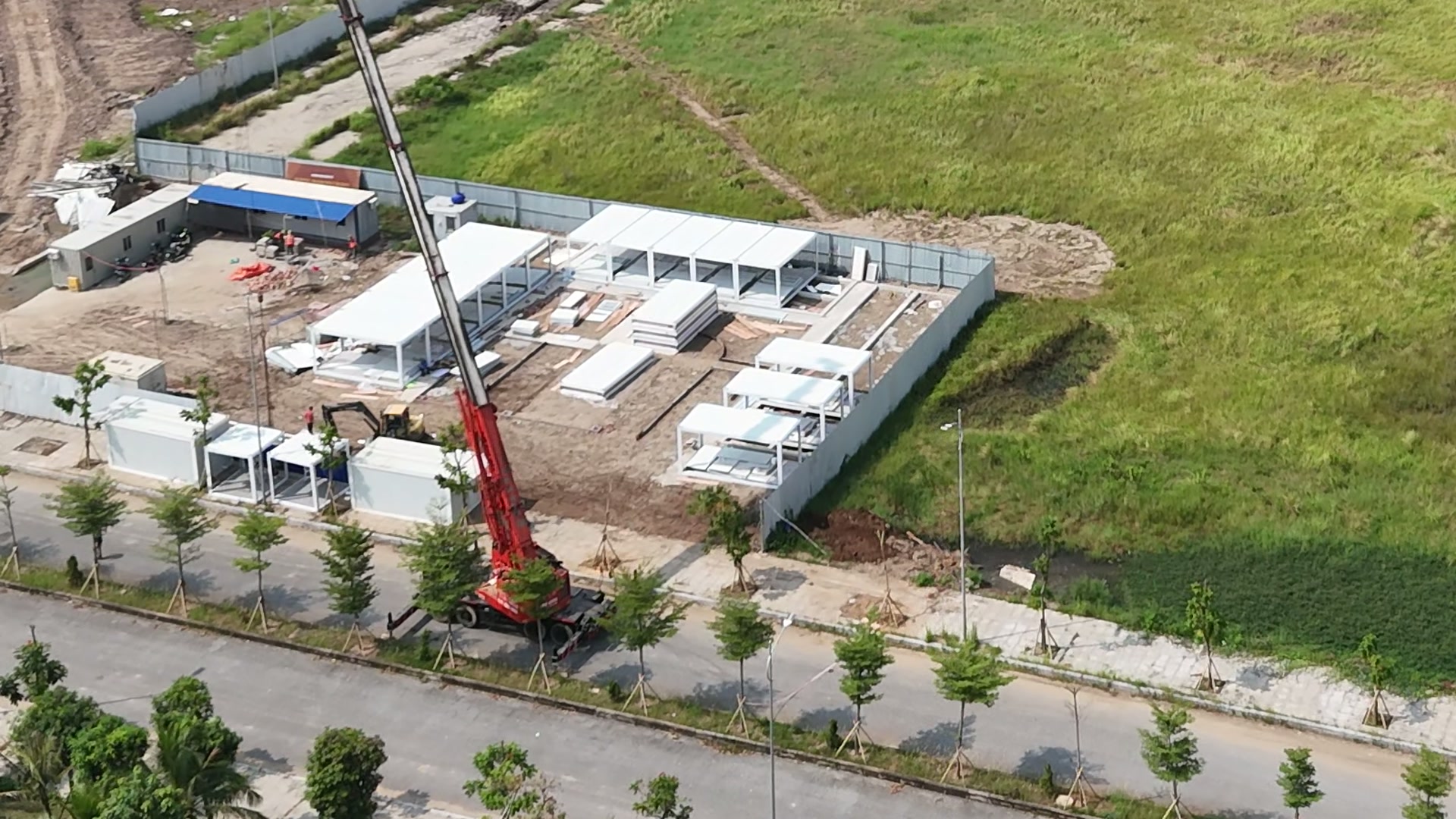 Khu điều hành ở góc công trường Doji 75 Complex, mấy dãy nhà lắp ghép màu trắng đang dựng, một lán mái xanh, cần cẩu đỏ đứng ngoài hàng rào sát vỉa hè, bên cạnh là bãi cỏ