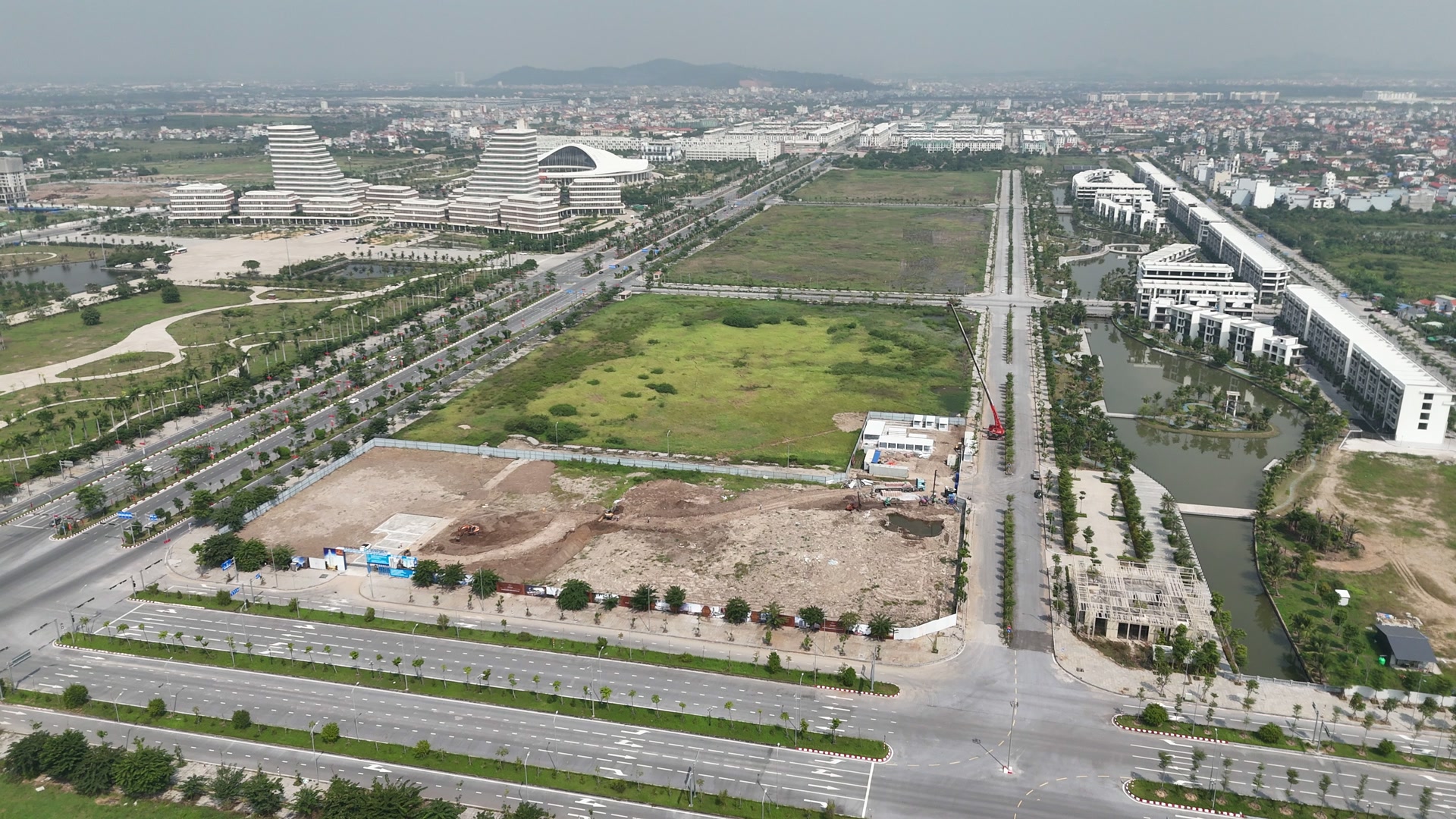 Lô đất Doji 75 Complex ở Bắc Sông Cấm quây tôn trắng nằm ở góc giao hai đường nhiều làn, mặt lô đã dọn, góc trong có khu nhà điều hành và cần cẩu đỏ, phía xa là khu Trung tâm hành chính mới