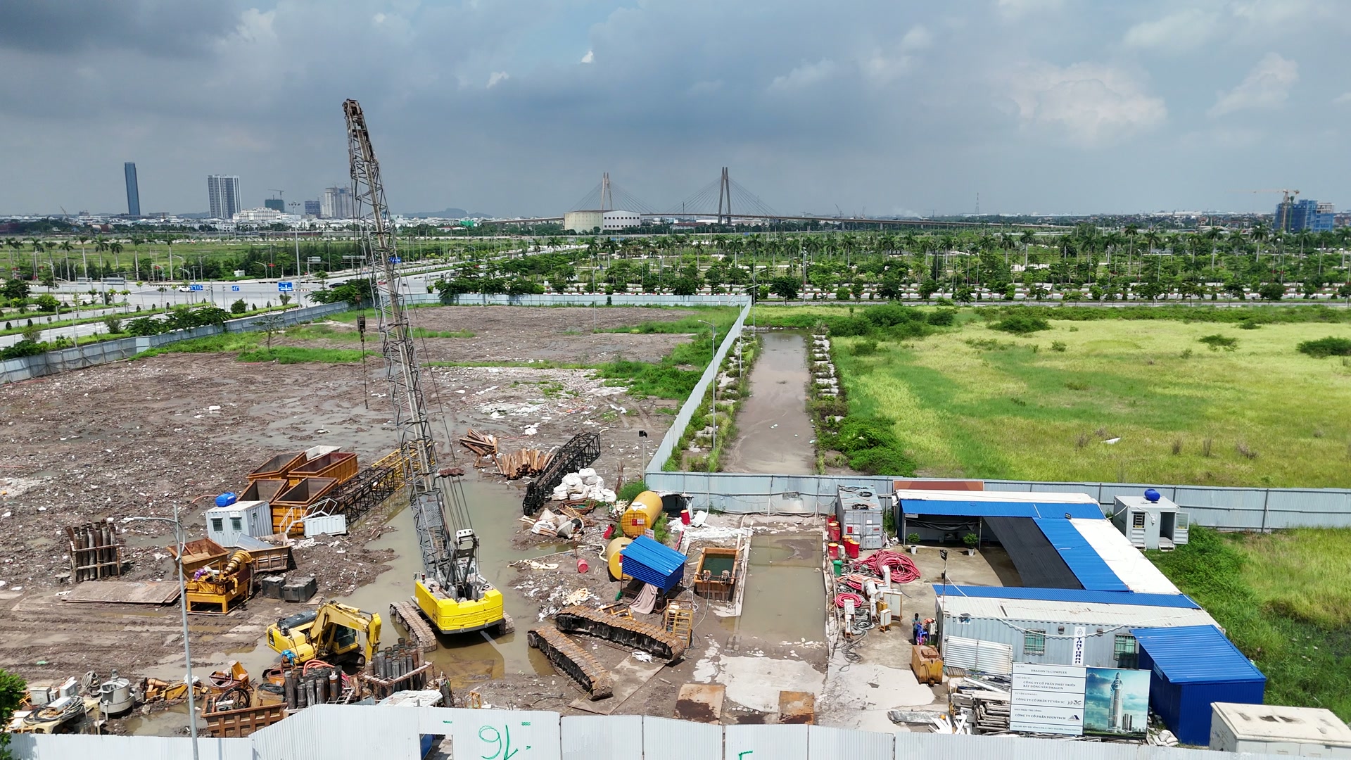 Lô Doji 75 Complex tháng 7/2026, mặt lô san phẳng còn đất đá và nước đọng, góc sát đường có cần cẩu bánh xích dựng cần, máy xúc vàng, lồng thép, container và lán mái xanh