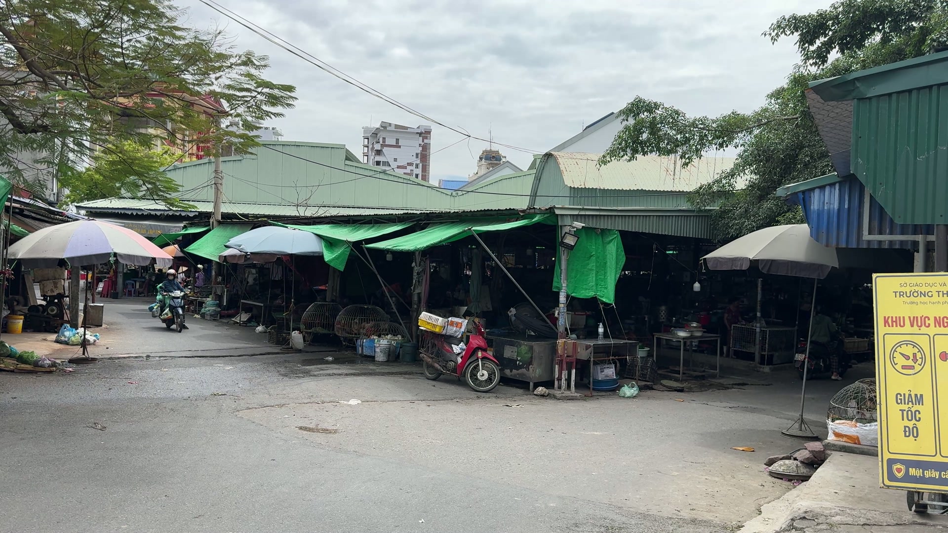 Dãy sạp chợ Núi Đèo mái tôn xanh, che ô nhiều màu, có người đi xe máy vào chợ và xe máy dựng trước sạp hàng
