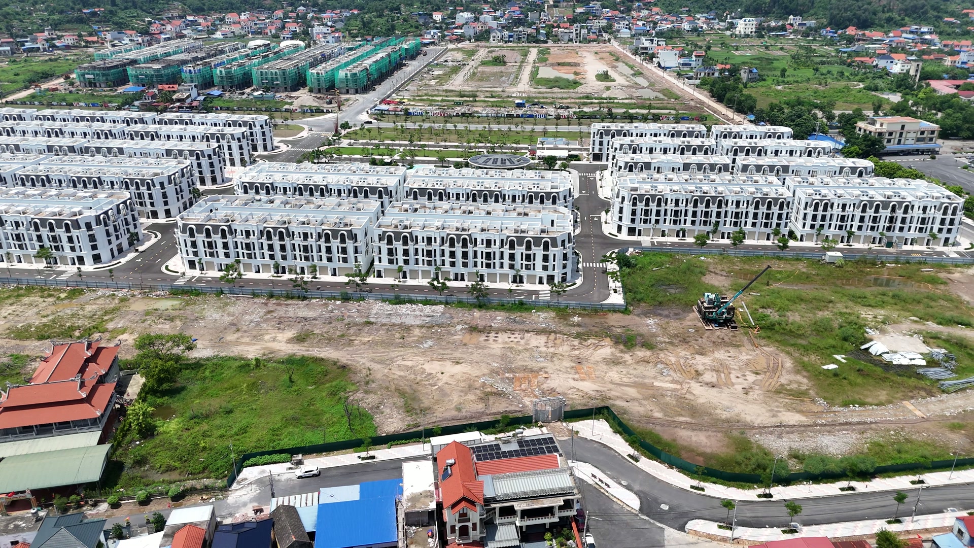 Khu đất còn trống trong dự án nhìn từ trên cao, có máy đào trên nền đất, phía sau là các dãy nhà thấp tầng màu trắng đã xây xong