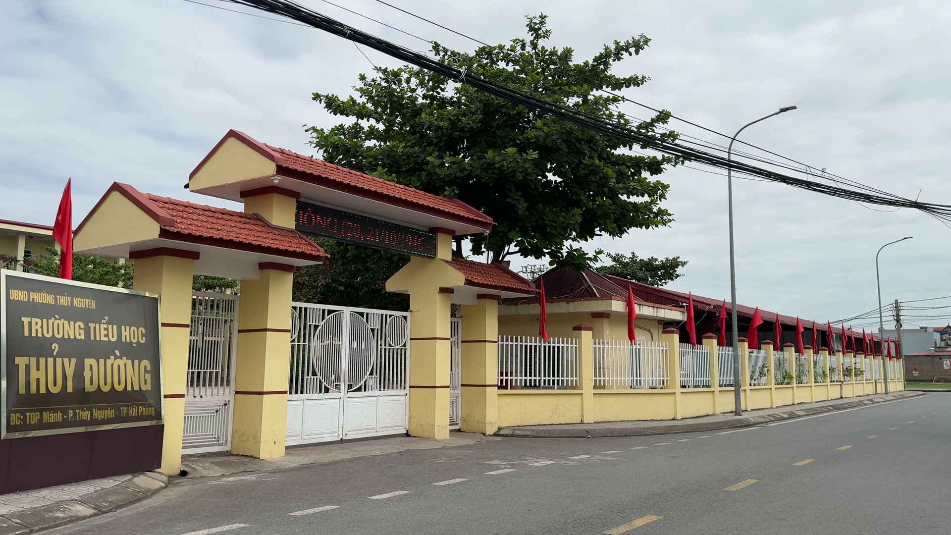 Cổng Trường Tiểu học Thủy Đường mái ngói đỏ, cột vàng, biển tên trường ở bên trái cổng, phía trước là con đường nhựa hai làn