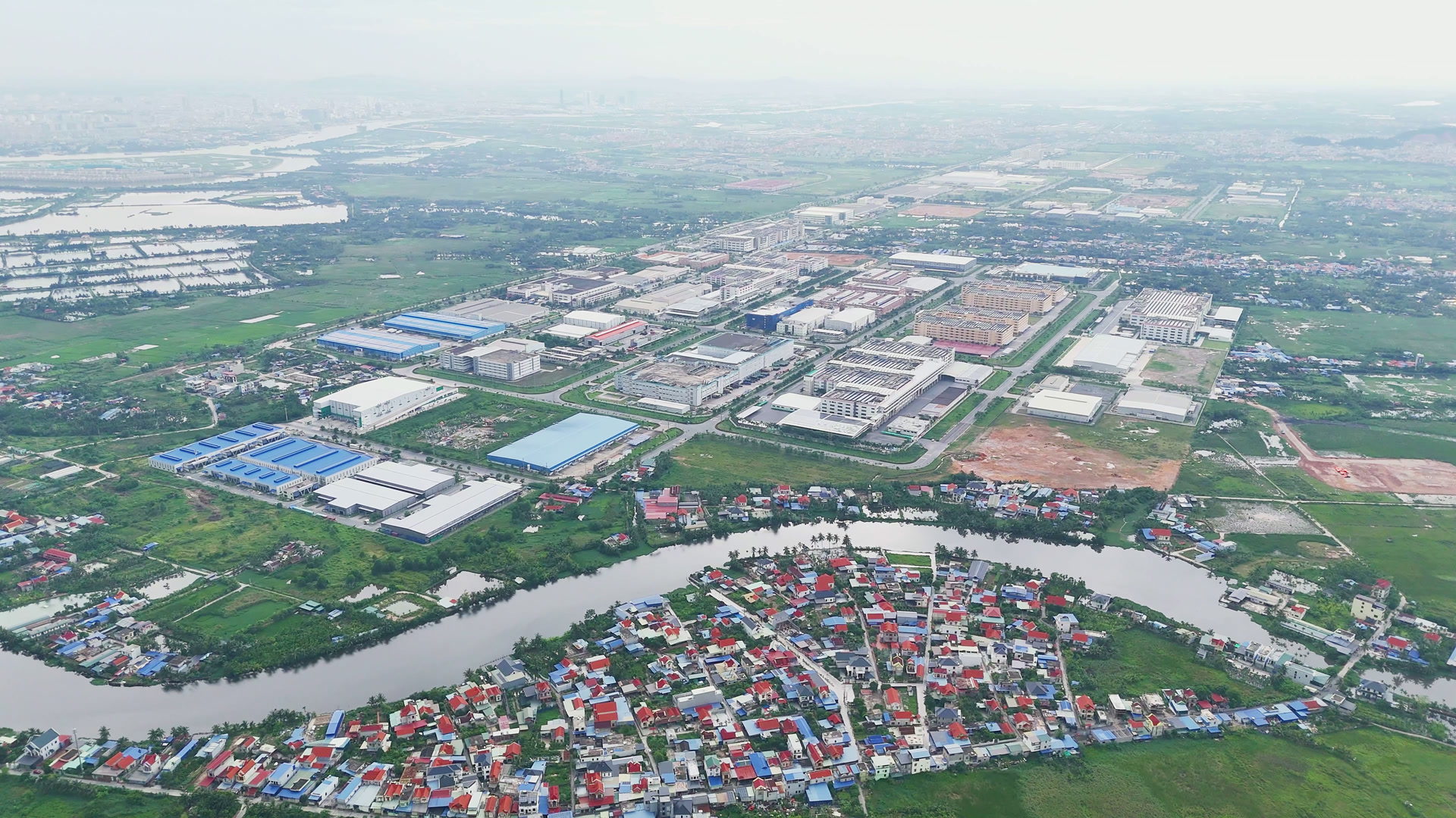 Những dãy nhà xưởng mái xanh và mái trắng trải dài trong một khu công nghiệp, bên dưới là một con sông nhỏ uốn quanh làng xóm nhà mái đỏ