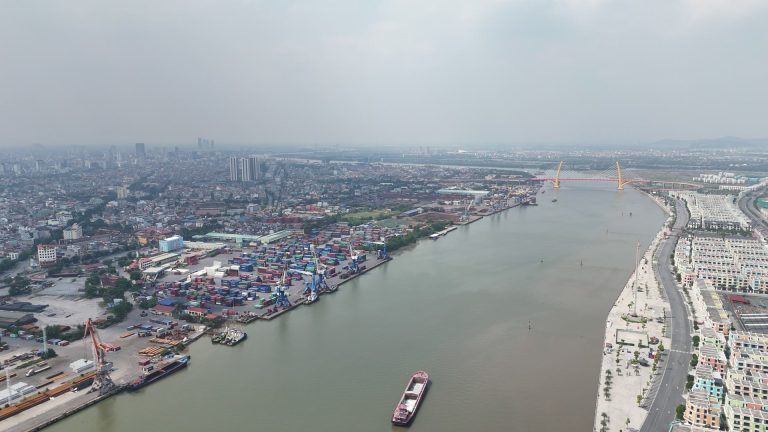 Toàn cảnh khúc sông Cấm, bên trái là dải bến đang hoạt động với bãi container và cần cẩu chân đế xanh, bên phải là dãy nhà phố trên đảo Vũ Yên, phía xa là cầu Hoàng Gia hai trụ tháp vàng
