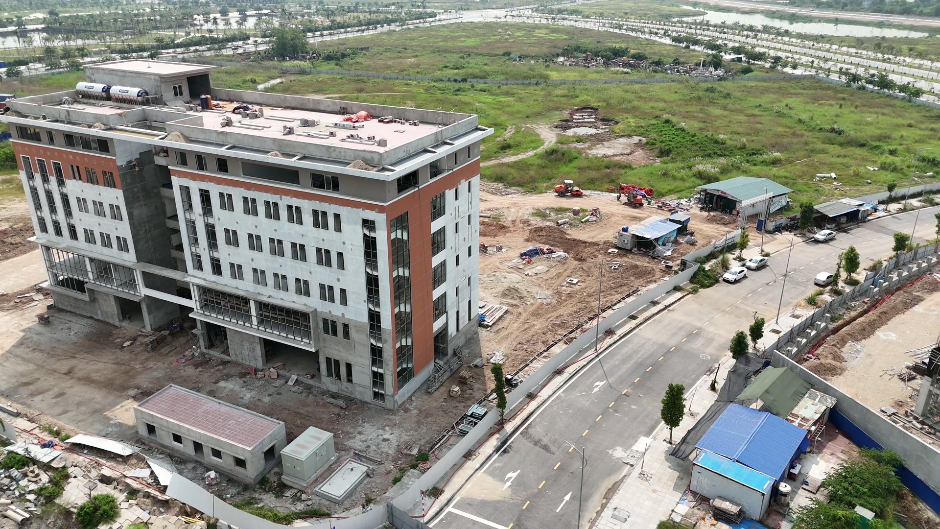 Toà nhà sáu tầng của tổ hợp giáo dục EQuest ở Bắc Sông Cấm nhìn chéo từ trên cao, mặt ngoài trắng có mảng nâu đỏ ở đầu hồi, quanh chân nhà là đất công trường, lán mái xanh và đường nội bộ cong