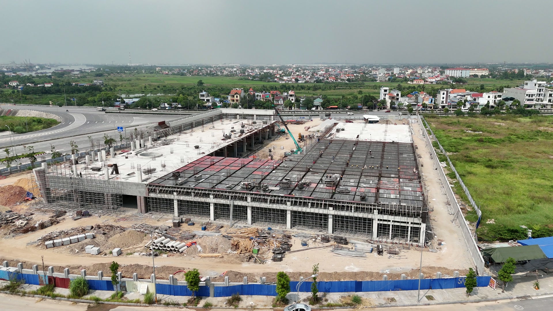 Công trường trung tâm thương mại chân cầu Bính, bên trái là mảng sàn bê tông đã làm xong còn thép chờ cột nhô lên, bên phải là mảng sàn đang dựng khuôn với nhiều thợ đứng trên mặt sàn và một chiếc cẩu màu xanh