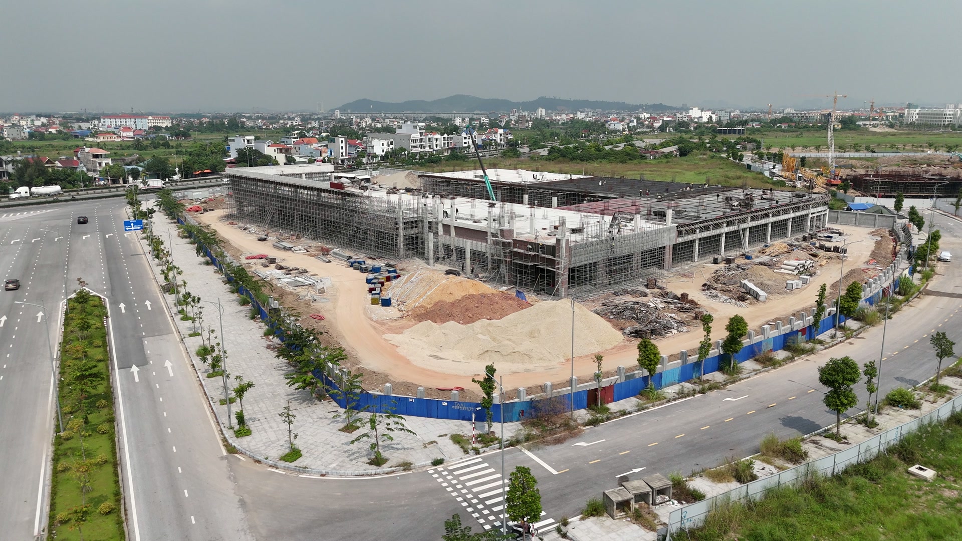Sân trước trung tâm thương mại chân cầu Bính vẫn là đất, cát và đá đổ thành đống lớn, toà nhà phía sau còn giàn giáo quây quanh, ngoài cùng là hàng rào tôn xanh và đường phố
