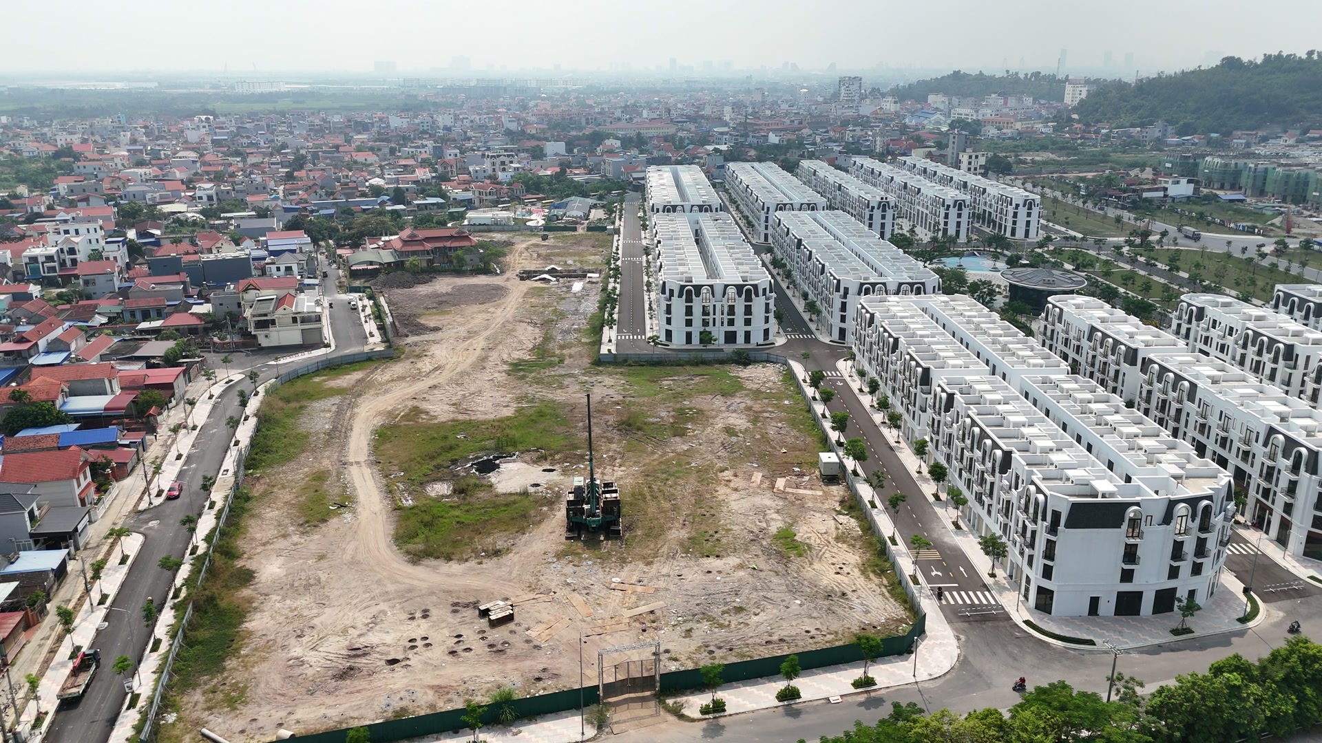 Lô đất trống ở Vlasta Thủy Nguyên nằm giữa làng xóm và các dãy nhà phố trắng đã hoàn thiện, giữa lô có một máy khoan cọc dựng cần, quanh đó là vệt bánh xe và đất mới đào, phần còn lại là cỏ
