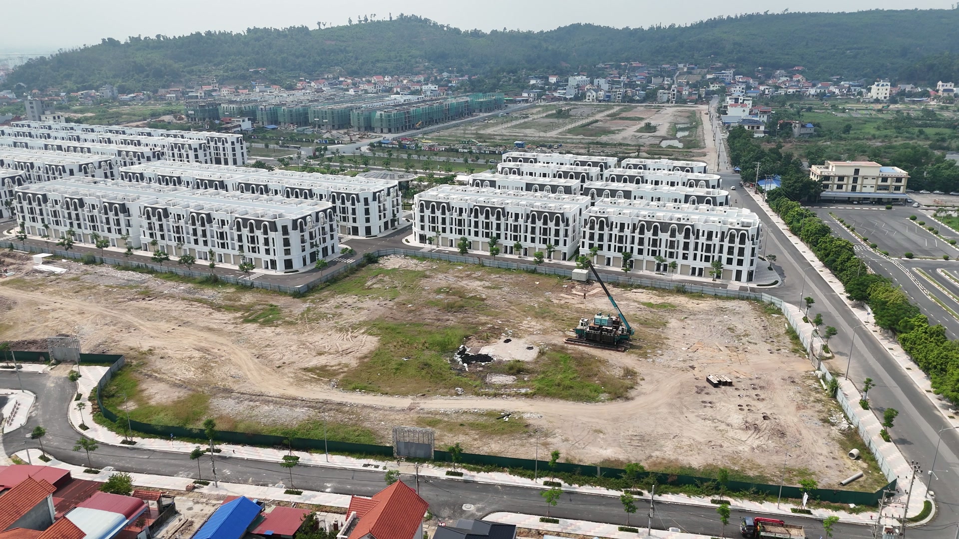 Lô đất trống rộng ở Vlasta Thủy Nguyên có hàng rào quây quanh, giữa lô là một máy khoan cọc dựng cần và vệt đất mới đào, phía sau là các dãy nhà phố trắng đã hoàn thiện