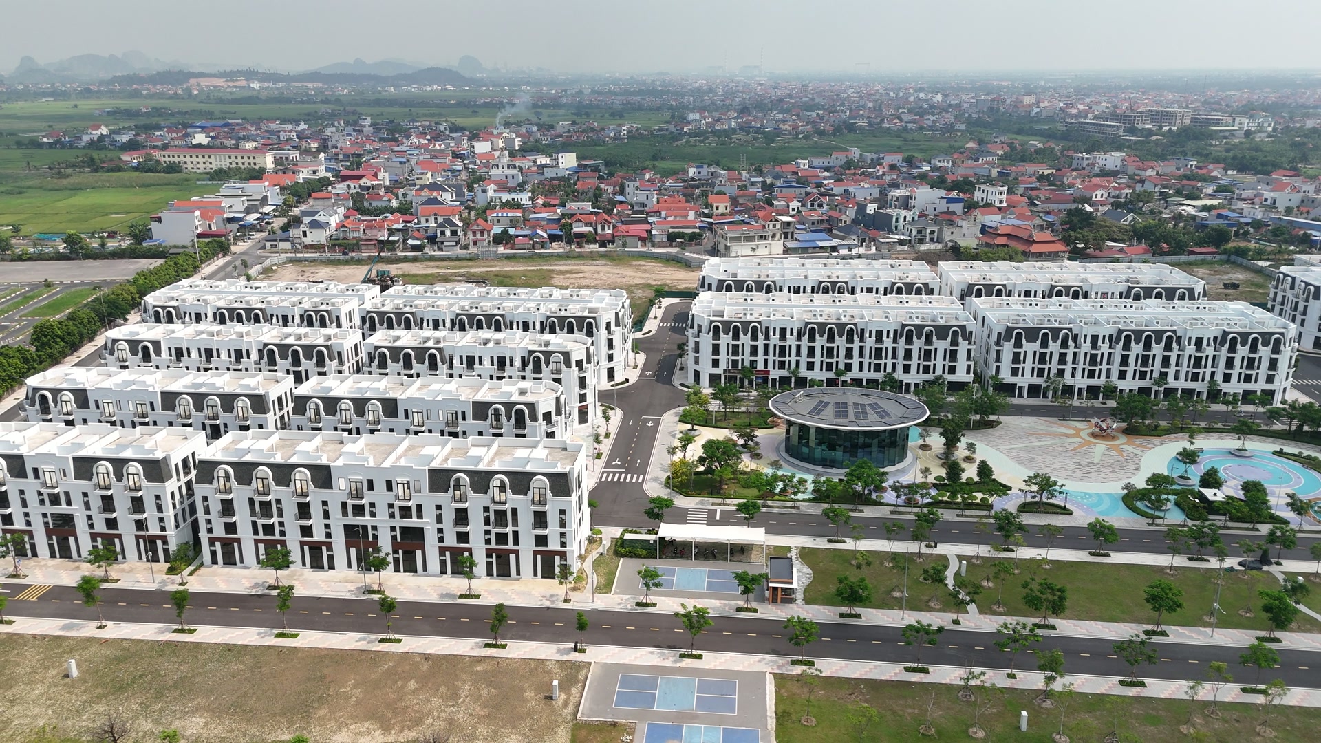 Các dãy nhà phố trắng đã hoàn thiện ở Vlasta Thủy Nguyên, giữa khung là công trình mái tròn lợp kính, quanh đó là khu cảnh quan có cây xanh và lối đi, phía trước là đường nhựa có hàng cây