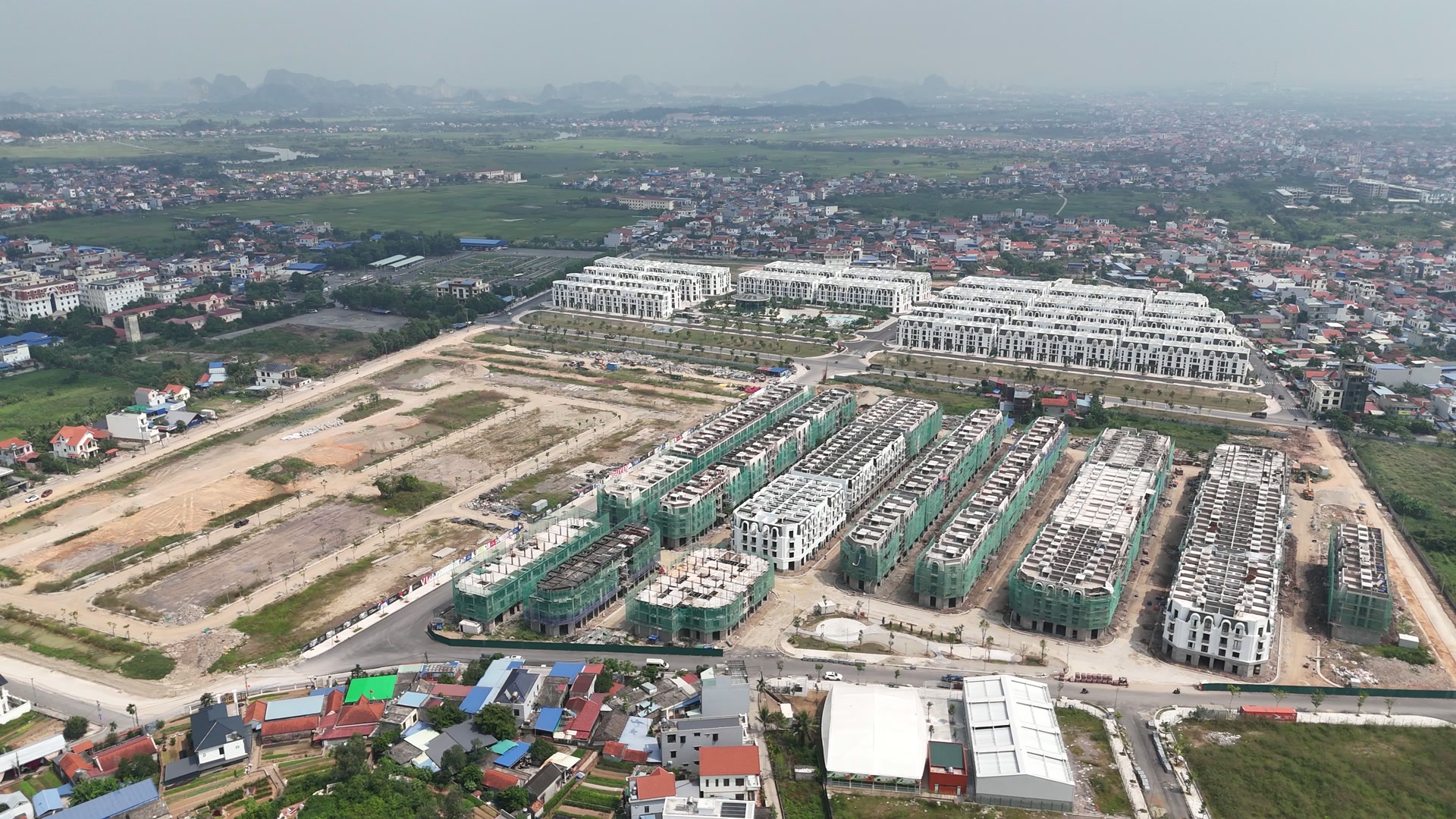 Nhìn chéo xuống các dãy nhà phố ở Vlasta Thủy Nguyên giữa tháng 10/2026, dãy đã tháo lưới tường trắng xen với dãy còn quây lưới xanh, phía sau là bãi đất đã chia ô, góc dưới là hai mái nhà lớn màu trắng và xám