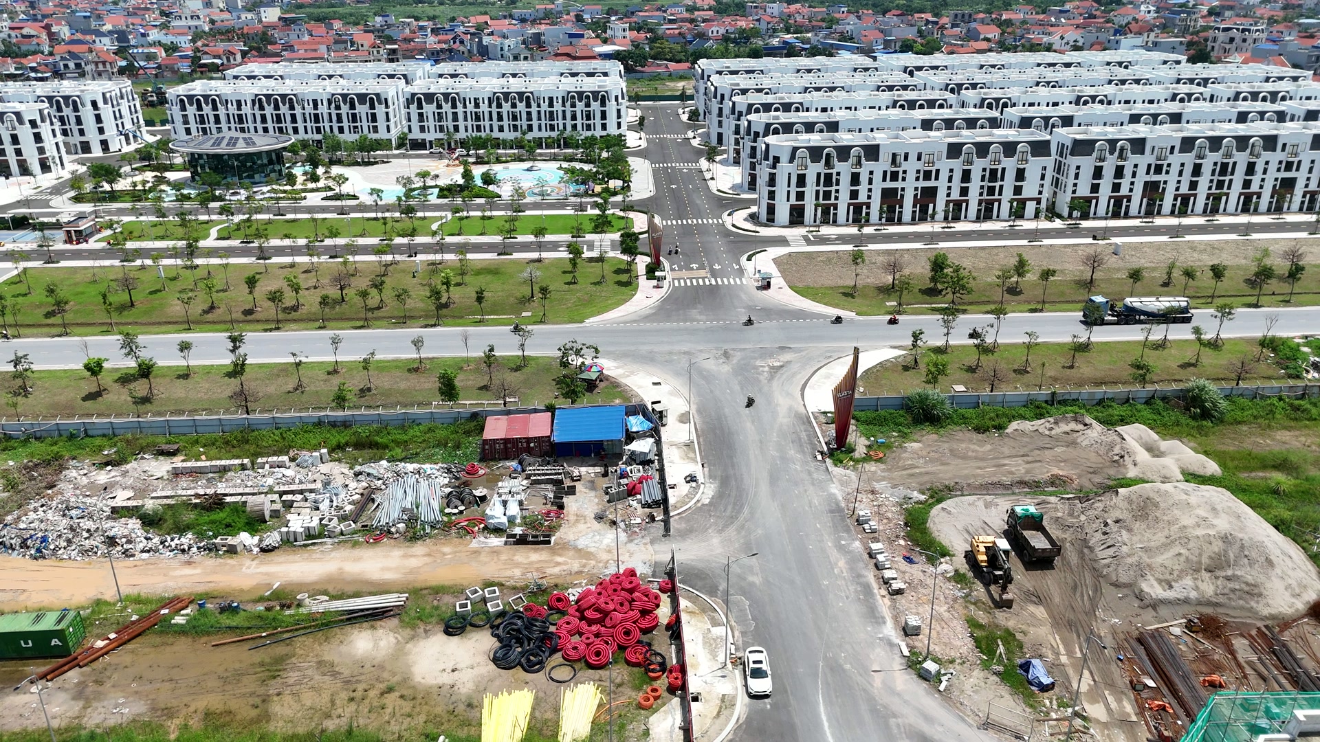 Nút giao trên trục đường 359C ở Vlasta Thủy Nguyên cuối tháng 8/2026, hai bên đường còn ngổn ngang đống cát đá, cuộn ống đỏ, máy xúc và lán mái xanh dựng tạm, phía xa là dãy nhà trắng