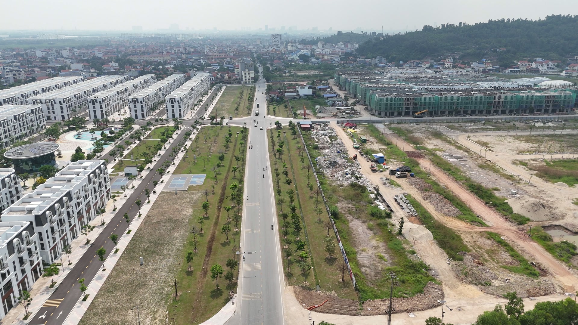 Trục đường 359C nhiều làn có dải cây giữa chạy thẳng vào khung, bên trái là dãy nhà phố trắng, bên phải là đất đã san phẳng chia thành từng ô, vật liệu và lán công trường để dọc mép đường